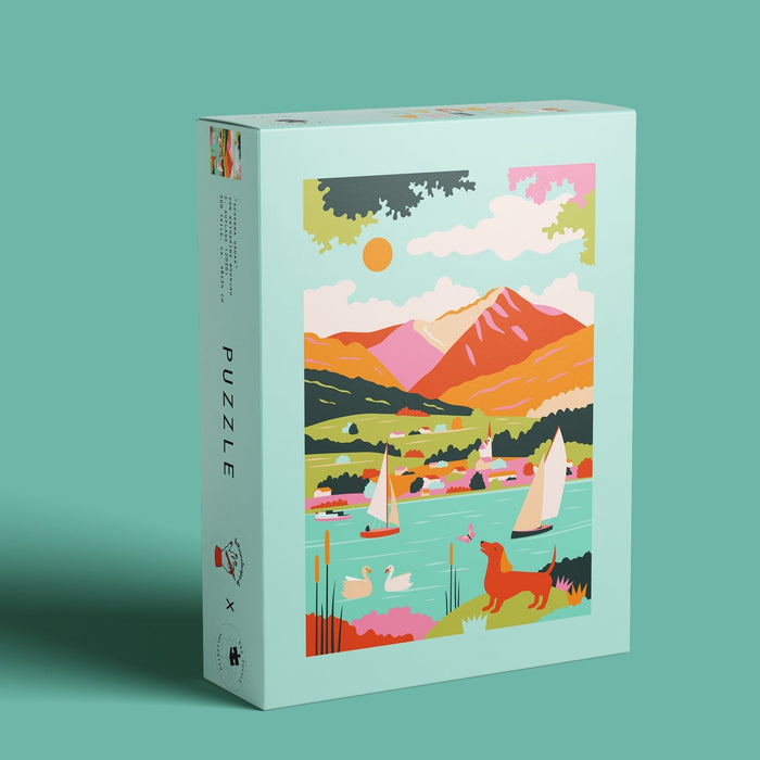 The Best Regards | 500 Piece Jigsaw Puzzle by Das Puzzle Kollektiv is a rectangular, plastic-free puzzle featuring colorful lake artwork with sailboats, swans, mountains, and a dog on a teal background.