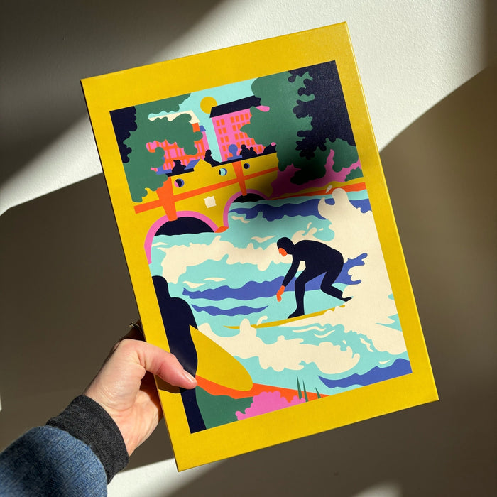 A hand holds Das Puzzle Kollektiv's Eisbach Wave | 1,000 Piece Jigsaw Puzzle, featuring a vibrant surfer scene with buildings, trees, and a bridge—plastic-free and climate-neutral, bordered by a yellow frame.