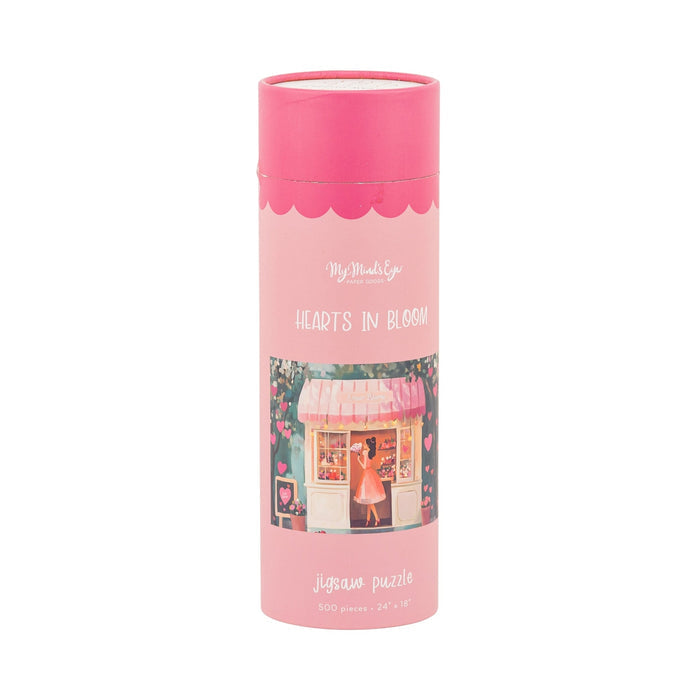 My Mind's Eye's Hearts in Bloom | 500 Piece Jigsaw Puzzle comes in a pink cylindrical container and features a charming flower shop illustration with a woman outside—an ideal romantic gift for Valentine's Day.